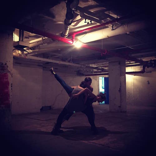 Two dancers perform in a dark, dingy basement. One dancer in arabesque while two dancers hold onto them.